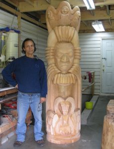 Photo of Ya'Ya Heit standing beside carved wooden sculpture with Indigenous west coast mythical representations of animals.