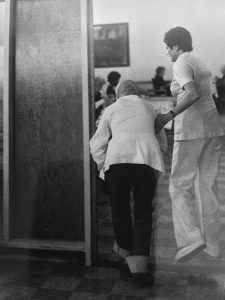 elderly woman walking through doorway with uniformed woman holding her arm