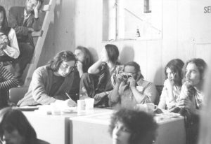 Group of early MPA members meeting around a long table. Lanny Beckman is writing at one end of the table, while Stan Persky, is holding a camera.